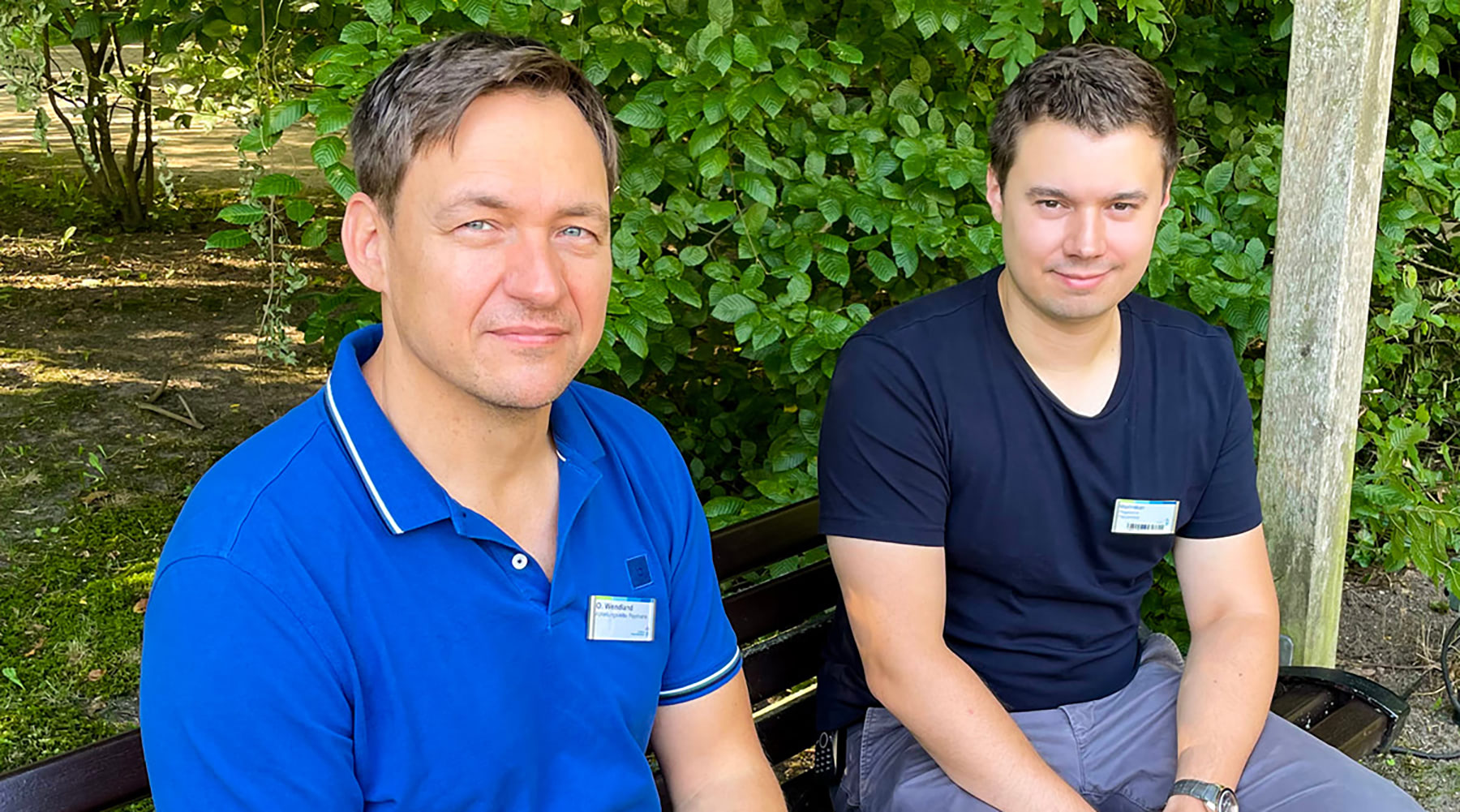 Oliver Faul-Wendland und Maxiliam Steckler sitzen nebeneinander auf einer Bank im Grünen und lächeln in die Kamera