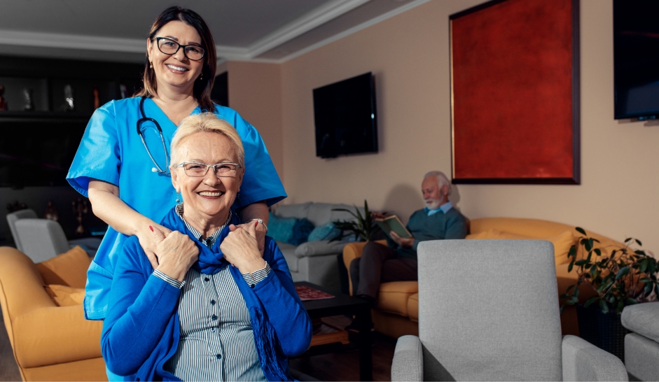 Eine Pflegekraft liegt ihre Hände auf die Schultern einer älteren Dame und beide lächeln in die Kamera