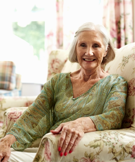 Eine ältere Dame sitzt in einem geblümten Sessel und lächelt in die Kamera